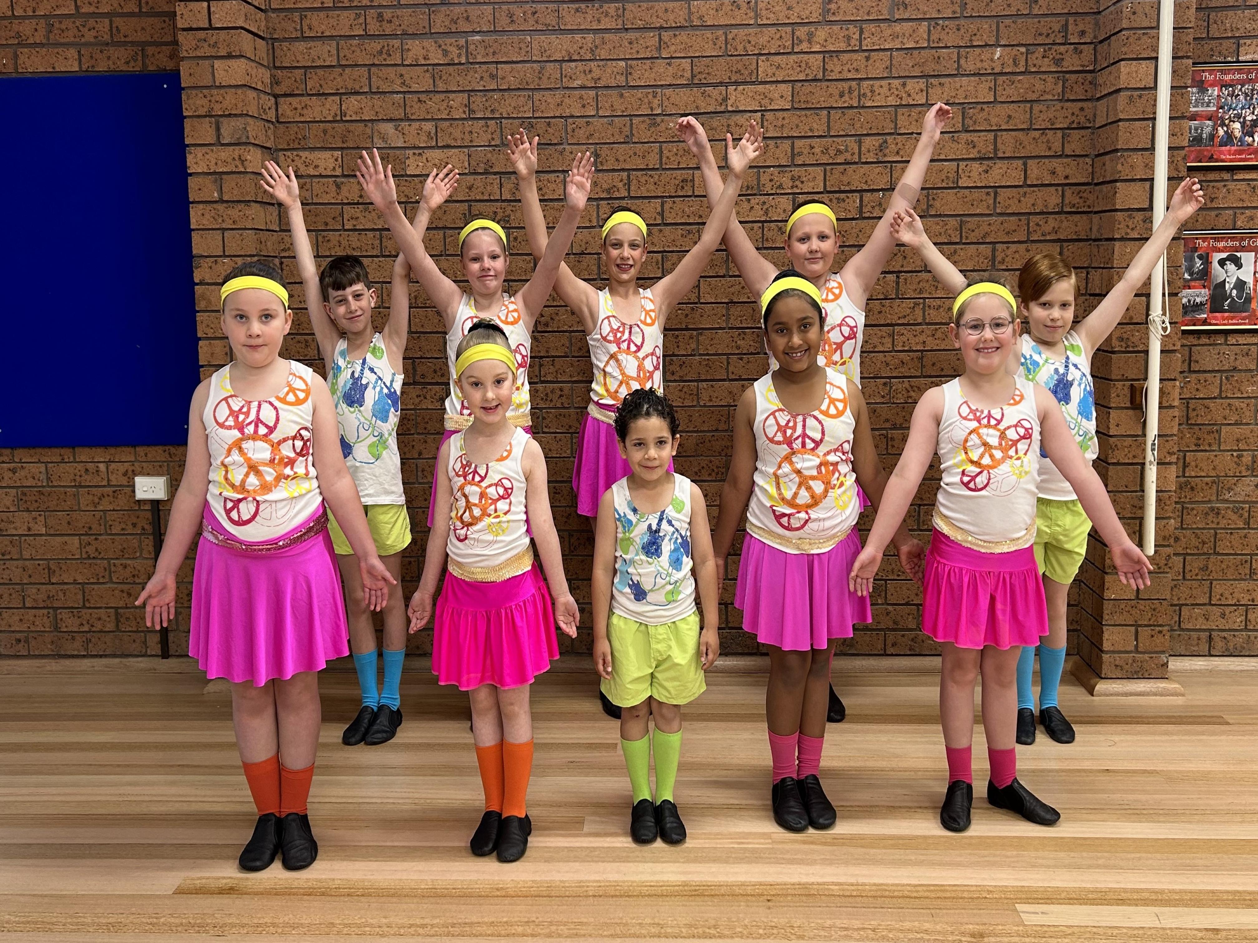 A group of very vibrant and colourful jazz and tap students wearing neon pink and neon yellow headbands, singlets, socks, and skirts.